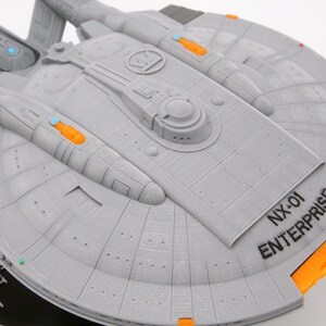 Puede incluir: Modelo gris de la nave espacial NX-01 Enterprise con detalles en naranja y azul. El modelo tiene el texto "NX-01 ENTERPRISE" impreso en el casco. El modelo est&aacute; montado sobre una base negra.