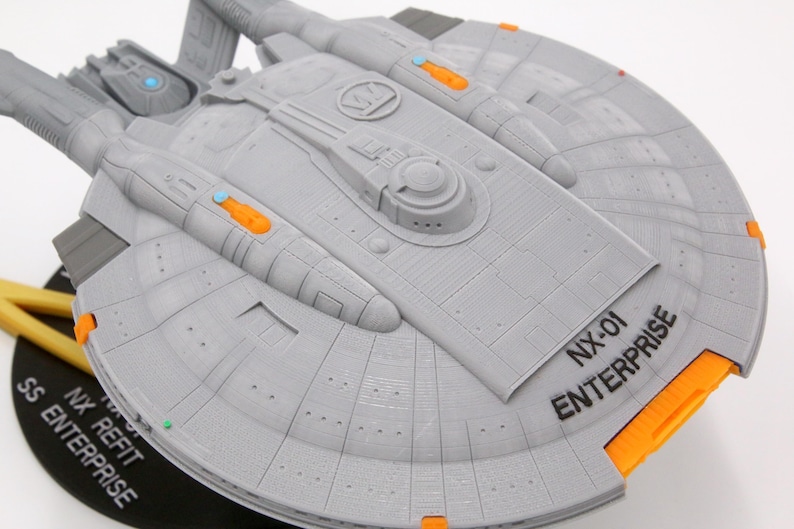 Puede incluir: Modelo gris de la nave espacial NX-01 Enterprise con detalles en naranja y azul. El modelo tiene el texto "NX-01 ENTERPRISE" impreso en el casco. El modelo est&aacute; montado sobre una base negra.