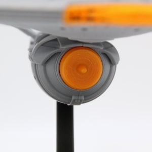 Puede incluir: Vista de cerca de una maqueta gris de una nave espacial con un componente de motor circular naranja. El modelo est&aacute; montado sobre un soporte negro sobre un fondo blanco. El dise&ntilde;o de la nave espacial es futurista.