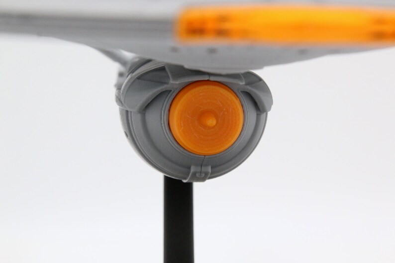 Puede incluir: Vista de cerca de una maqueta gris de una nave espacial con un componente de motor circular naranja. El modelo est&aacute; montado sobre un soporte negro sobre un fondo blanco. El dise&ntilde;o de la nave espacial es futurista.