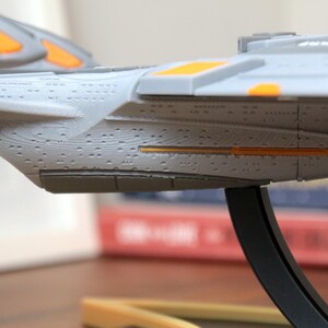 Puede incluir: Un modelo gris y naranja de una nave espacial con una textura de superficie detallada. La nave espacial est&aacute; montada en un soporte negro.