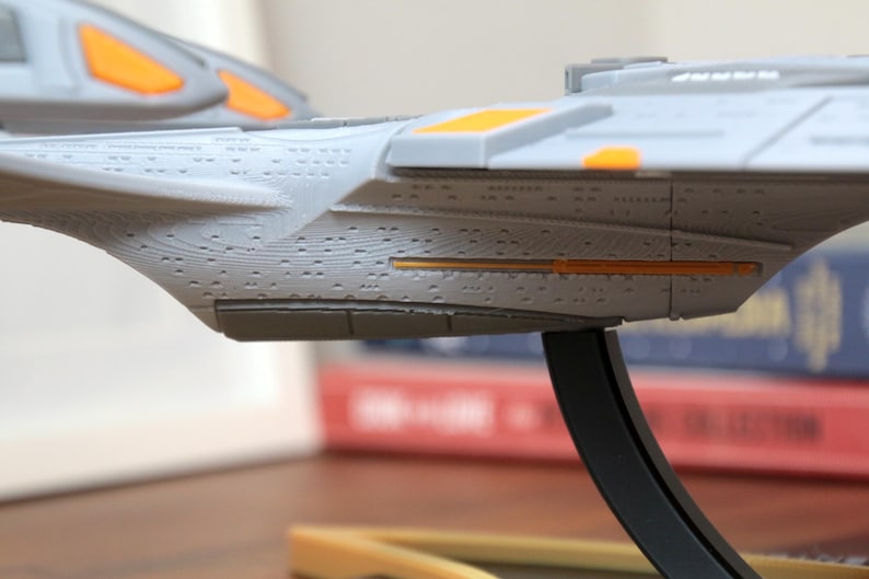 Puede incluir: Un modelo gris y naranja de una nave espacial con una textura de superficie detallada. La nave espacial est&aacute; montada en un soporte negro.