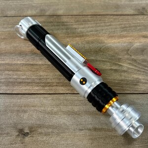Puede incluir: Un accesorio de sable de luz plateado, negro y dorado con un bot&oacute;n rojo y dos puntos amarillos. El accesorio est&aacute; sobre una superficie de madera. El sable de luz tiene una empu&ntilde;adura plateada y un agarre negro.
