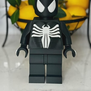 May include: A black and white figurine resembling a superhero, with a distinctive white spider emblem on the chest and white eye shapes. The figurine is made of interlocking blocks, similar to a popular building toy.
