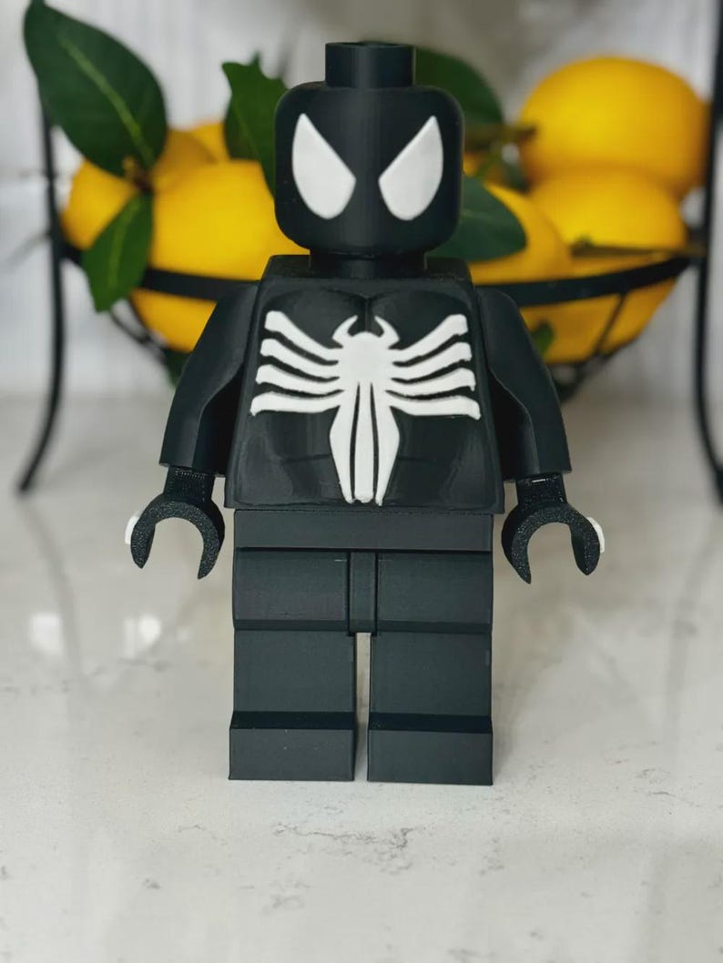 May include: A black and white figurine resembling a superhero, with a distinctive white spider emblem on the chest and white eye shapes. The figurine is made of interlocking blocks, similar to a popular building toy.