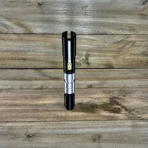 May include: A black and silver lightsaber prop with a yellow button and a black handle. The lightsaber is standing upright on a wooden surface. The top of the lightsaber has a vertical slit.