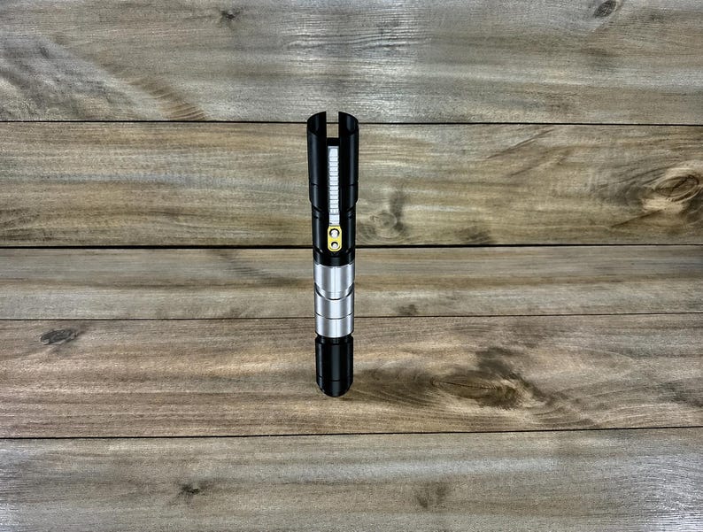 May include: A black and silver lightsaber prop with a yellow button and a black handle. The lightsaber is standing upright on a wooden surface. The top of the lightsaber has a vertical slit.