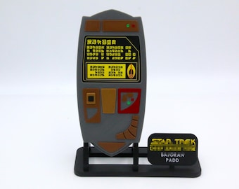 May include: A gray and brown Star Trek prop, a Bajoran PADD, with a black base. The PADD features yellow and green accents, with text in an alien language. The base has the words "STAR TREK" and "BAJORAN PADD".