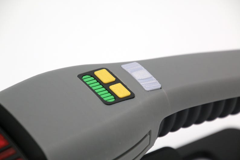 K&ouml;nnte beinhalten: Ein graues Handger&auml;t mit einem schwarzen Panel, auf dem sich ein gr&uuml;ner und gelber Knopf befindet. Das Ger&auml;t hat einen strukturierten schwarzen Griff.