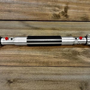 May include: A silver lightsaber with a black grip and red buttons. The lightsaber has a curved blade on each end. The lightsaber is lying on a wooden surface.