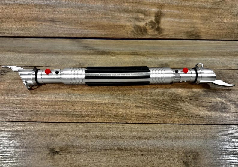 May include: A silver lightsaber with a black grip and red buttons. The lightsaber has a curved blade on each end. The lightsaber is lying on a wooden surface.