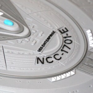 Puede incluir: Primer plano de una superficie circular gris texturizada con el texto "NCC-1701-E" y "USS Enterprise" impreso en negro.