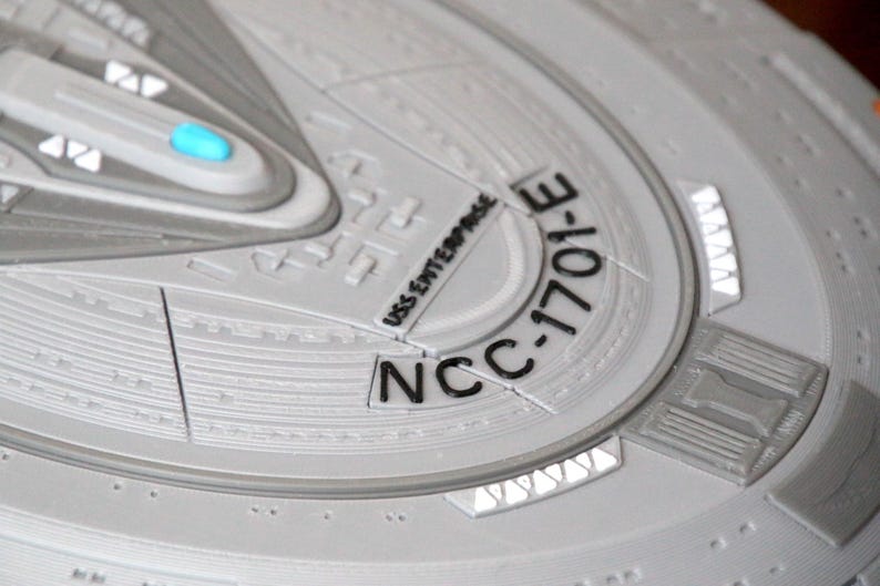 Puede incluir: Primer plano de una superficie circular gris texturizada con el texto "NCC-1701-E" y "USS Enterprise" impreso en negro.