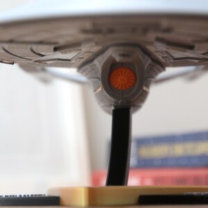 Puede incluir: Un modelo gris y plateado de una nave espacial con una luz naranja en la parte inferior. La nave espacial est&aacute; en un soporte negro con una base dorada.