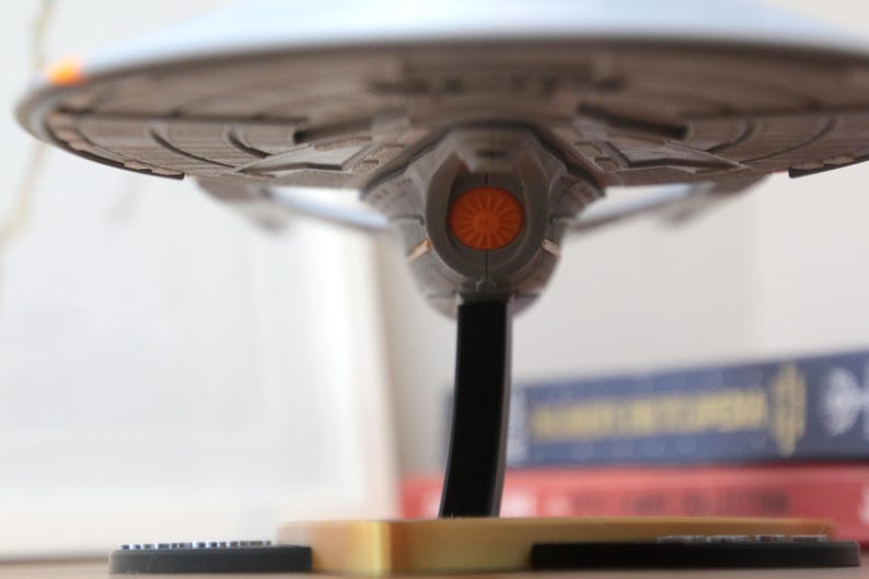 Puede incluir: Un modelo gris y plateado de una nave espacial con una luz naranja en la parte inferior. La nave espacial est&aacute; en un soporte negro con una base dorada.