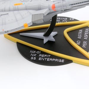 Puede incluir: Un modelo de la nave espacial NX-01 SS Enterprise, en gris y naranja, montado sobre una base negra con detalles amarillos. La base presenta el texto "NX-01 NX REFIT SS ENTERPRISE" y "AD ASTRA PER ASPERA".