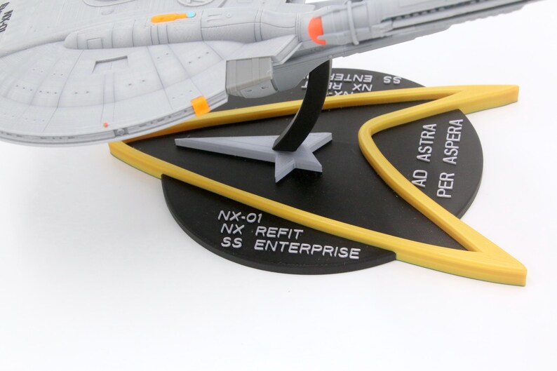 Puede incluir: Un modelo de la nave espacial NX-01 SS Enterprise, en gris y naranja, montado sobre una base negra con detalles amarillos. La base presenta el texto "NX-01 NX REFIT SS ENTERPRISE" y "AD ASTRA PER ASPERA".