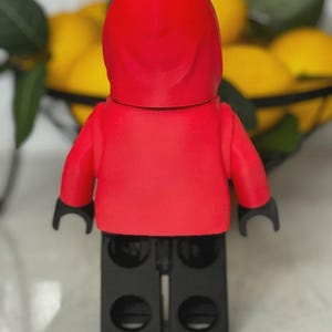 May include: A red minifigure with a hooded top and black arms and legs. The figure is standing in front of a blurred background of lemons in a black wire basket. The figure is a toy.
