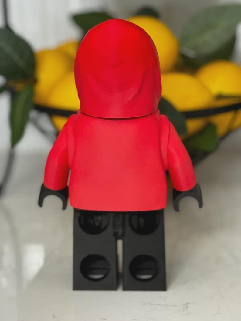May include: A red minifigure with a hooded top and black arms and legs. The figure is standing in front of a blurred background of lemons in a black wire basket. The figure is a toy.