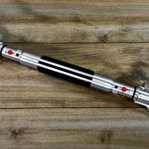 May include: A silver lightsaber with a black grip and red buttons. The hilt has a ribbed design and two curved blades extend from the top. The lightsaber is lying on a wooden surface.