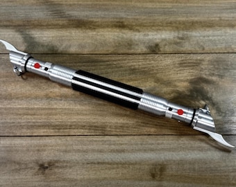 May include: A silver lightsaber with a black grip and red buttons. The hilt has a ribbed design and two curved blades extend from the top. The lightsaber is lying on a wooden surface.