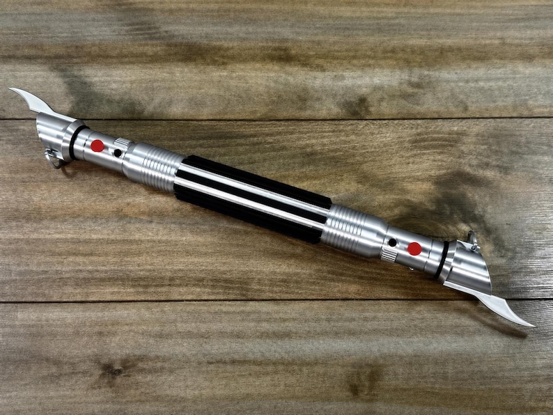 May include: A silver lightsaber with a black grip and red buttons. The hilt has a ribbed design and two curved blades extend from the top. The lightsaber is lying on a wooden surface.