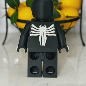 May include: A black Lego-style figure with a white spider emblem on the chest. The figure has a block-shaped head and arms. The background includes a wire basket filled with yellow lemons.
