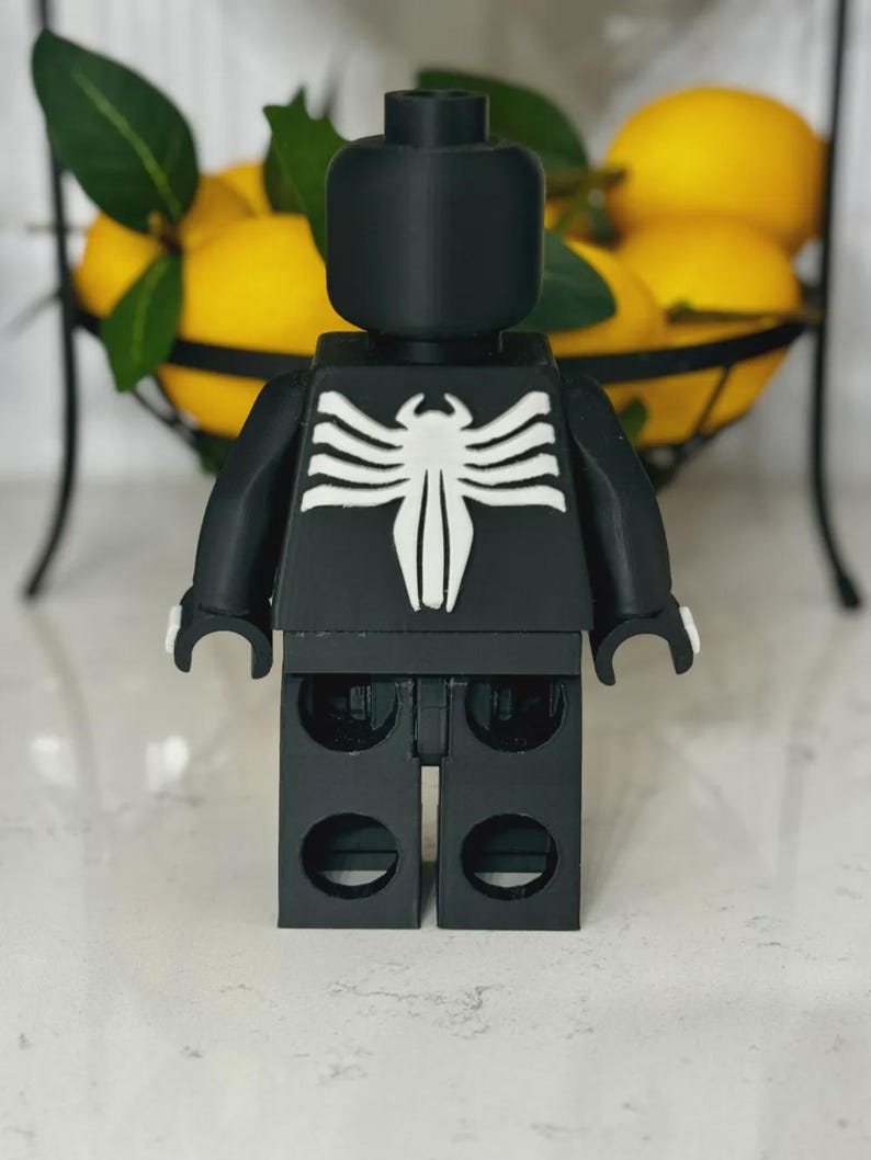 May include: A black Lego-style figure with a white spider emblem on the chest. The figure has a block-shaped head and arms. The background includes a wire basket filled with yellow lemons.