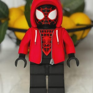 May include: A red and black Spider-Man figure wearing a red hoodie with white drawstrings. The figure has a black body, black legs, and black hands. The Spider-Man mask has white eyes and a red spiderweb design.