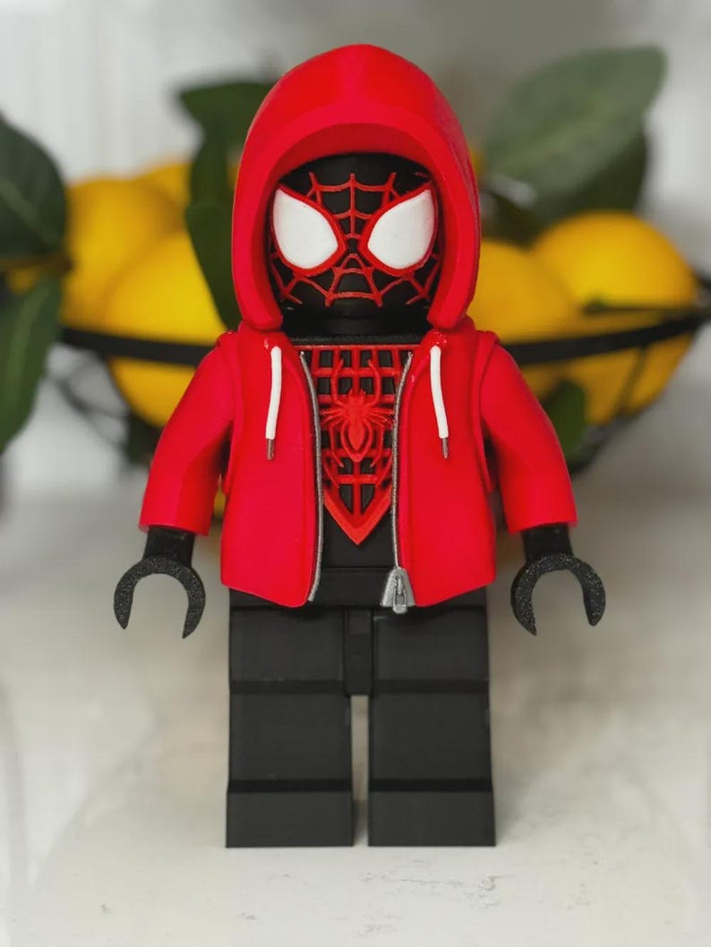 May include: A red and black Spider-Man figure wearing a red hoodie with white drawstrings. The figure has a black body, black legs, and black hands. The Spider-Man mask has white eyes and a red spiderweb design.