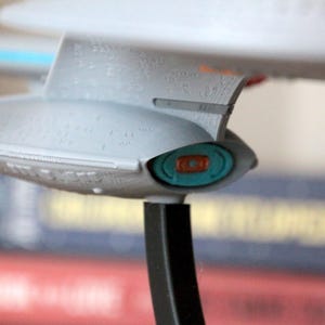 K&ouml;nnte beinhalten: Ein graues und wei&szlig;es Modell eines Raumschiffs mit einem blauen und orangefarbenen Detail. Das Raumschiff steht auf einem schwarzen St&auml;nder.