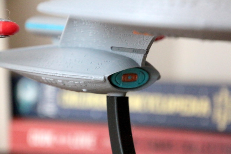 K&ouml;nnte beinhalten: Ein graues und wei&szlig;es Modell eines Raumschiffs mit einem blauen und orangefarbenen Detail. Das Raumschiff steht auf einem schwarzen St&auml;nder.