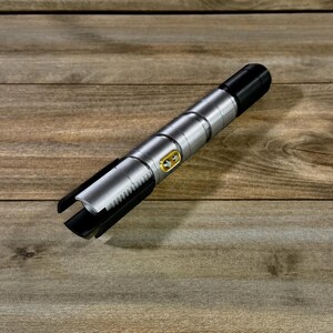 May include: A silver and black lightsaber prop with a gold button detail. The lightsaber is lying on a wooden surface. The lightsaber has a black handle and a silver body with a ribbed design.
