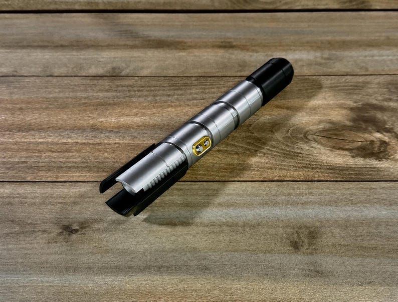 May include: A silver and black lightsaber prop with a gold button detail. The lightsaber is lying on a wooden surface. The lightsaber has a black handle and a silver body with a ribbed design.