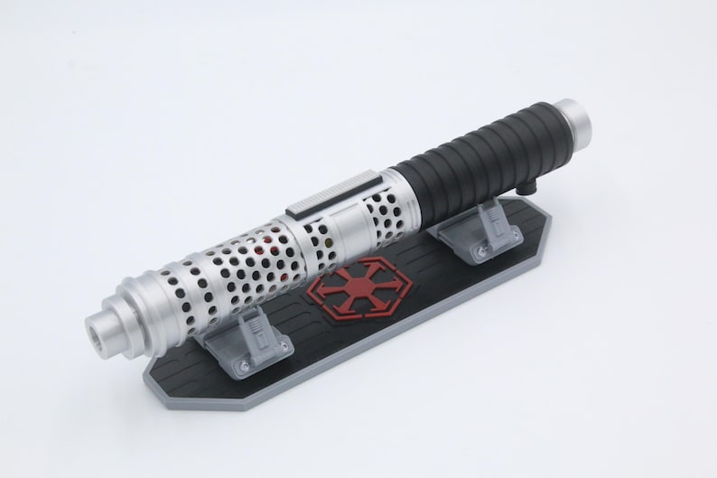 May include: A silver and black cylindrical lightsaber hilt featuring a perforated metal shroud, resting horizontally on a black and gray display stand that displays a red six-pointed emblem with arrows pointing outward from the center.