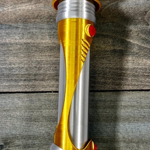 May include: A gold and silver lightsaber prop with red accents. The hilt is primarily silver with gold detailing, including a curved design and a circular top. Two red buttons are visible.