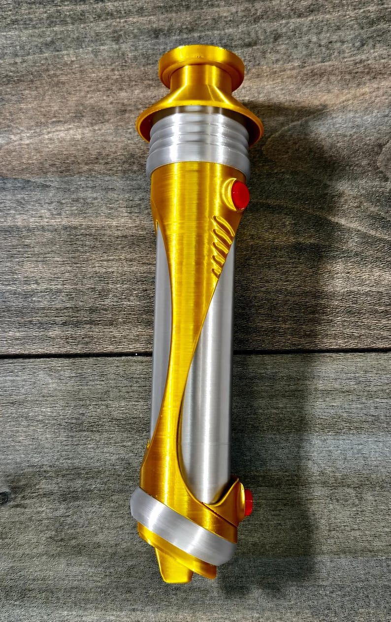 May include: A gold and silver lightsaber prop with red accents. The hilt is primarily silver with gold detailing, including a curved design and a circular top. Two red buttons are visible.