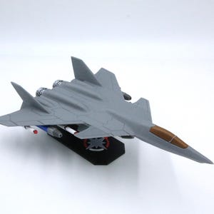 Può includere: Un modello di aereo grigio con un abitacolo marrone e componenti del motore dettagliati. L'aereo giocattolo ha un design elegante con ali e coda ed è montato su una base nera. Presenta accenti rossi, bianchi e blu sul lato inferiore.