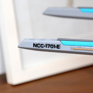 Puede incluir: Primer plano de una maqueta de nave espacial blanca con una franja azul y naranja. La nave espacial tiene el texto "NCC-1701-E" impreso en el lateral.