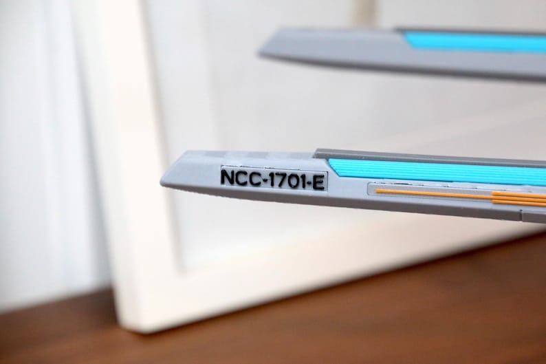 Puede incluir: Primer plano de una maqueta de nave espacial blanca con una franja azul y naranja. La nave espacial tiene el texto "NCC-1701-E" impreso en el lateral.