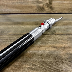 May include: A silver and black lightsaber prop with a red button and a blade-like emitter. The prop is made of metal and plastic, and it is lying on a wooden surface.