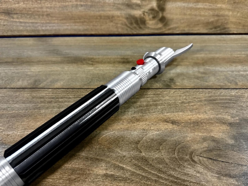 May include: A silver and black lightsaber prop with a red button and a blade-like emitter. The prop is made of metal and plastic, and it is lying on a wooden surface.