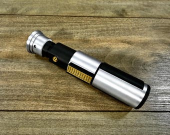 May include: A silver, black, and gold lightsaber prop, lying on a wooden surface. The prop features a metallic silver handle, a black midsection, and gold detailing. The lightsaber is a replica of a popular science fiction weapon.