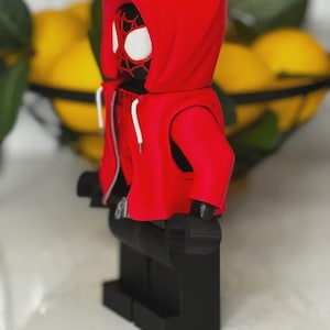 May include: A red hooded figure, resembling a superhero, stands on a black base. The figure has a red hoodie with white drawstrings and a black face with white eye details. The figure is a toy or collectible.