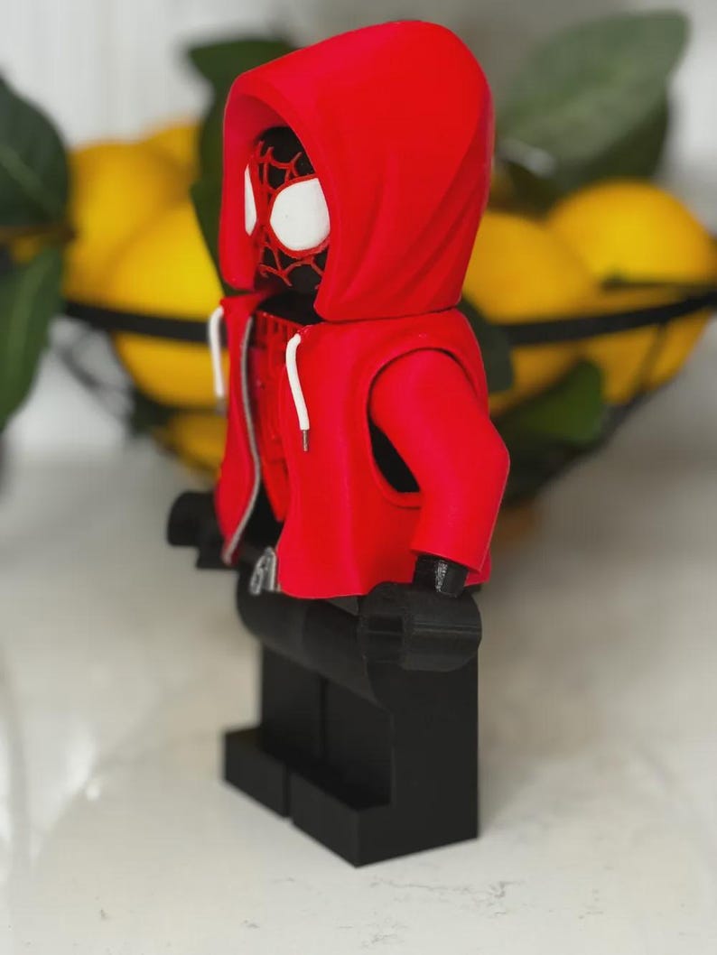 May include: A red hooded figure, resembling a superhero, stands on a black base. The figure has a red hoodie with white drawstrings and a black face with white eye details. The figure is a toy or collectible.