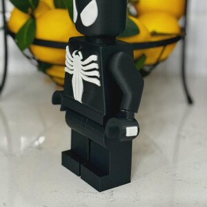 May include: A black and white Spider-Man-themed Lego figure. The figure has white eye shapes and a white spider emblem on the chest. The figure is standing on a white surface, with a wire basket of lemons in the background.