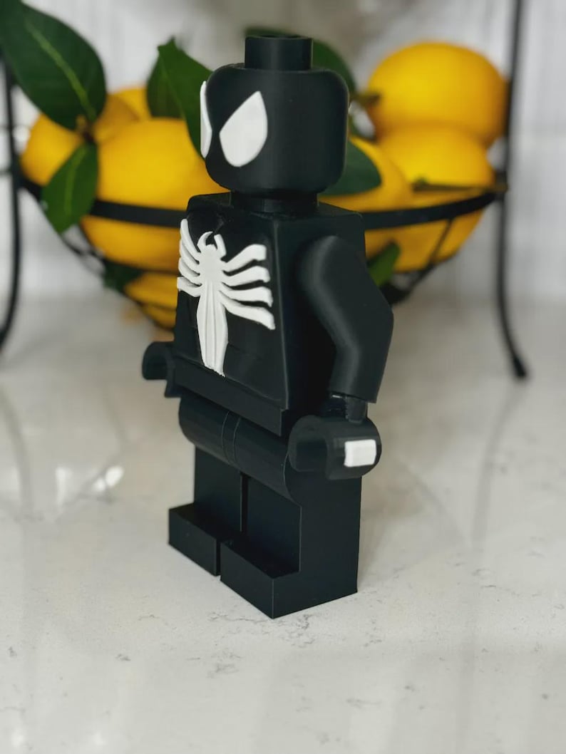 May include: A black and white Spider-Man-themed Lego figure. The figure has white eye shapes and a white spider emblem on the chest. The figure is standing on a white surface, with a wire basket of lemons in the background.