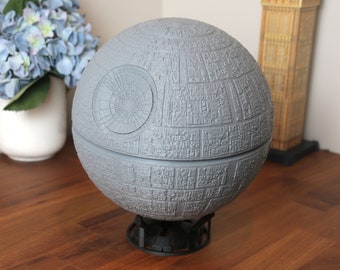 May include: A gray, spherical Death Star model with intricate details and a textured surface. The model is mounted on a black stand.