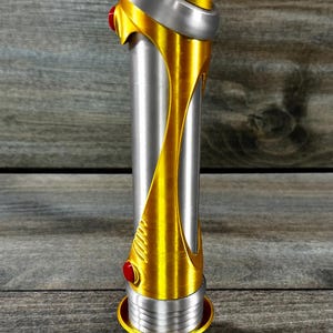 May include: A metallic gold and silver lightsaber with red accents. The hilt is cylindrical with a gold design that wraps around the silver body. The top and bottom have gold and silver details.
