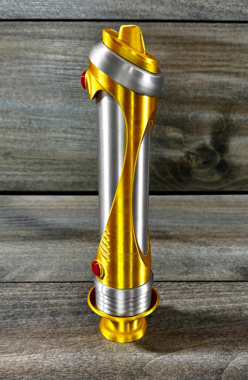May include: A metallic gold and silver lightsaber with red accents. The hilt is cylindrical with a gold design that wraps around the silver body. The top and bottom have gold and silver details.